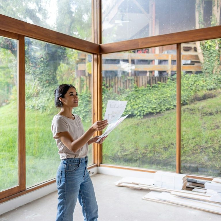 Femme tenant des plans, debout dans un espace lumineux avec de grandes fenêtres.